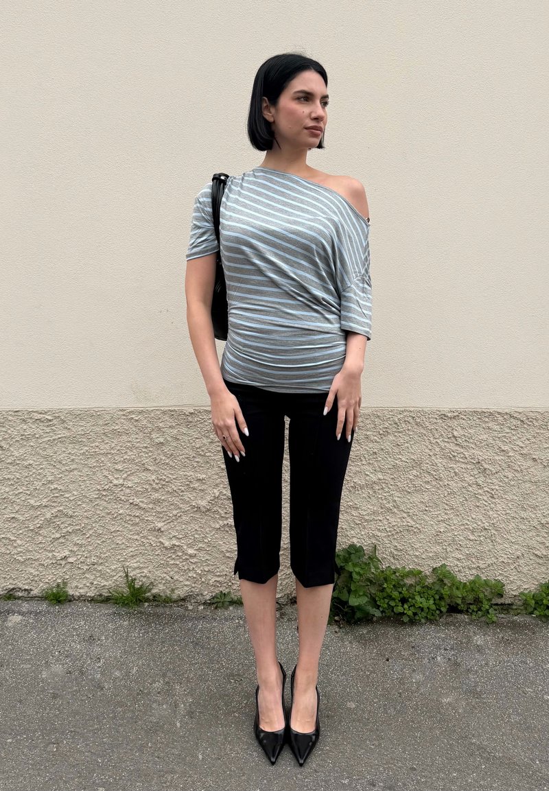 Γυναίκα με κοντά μαύρα μαλλιά στέκεται μπροστά σε μπεζ τοίχο, φορώντας ριγέ τοπάκι με ακάλυπτους τους ώμους, μαύρο κοντό παντελόνι, μαύρες γόβες και κρατώντας μαύρη τσάντα.