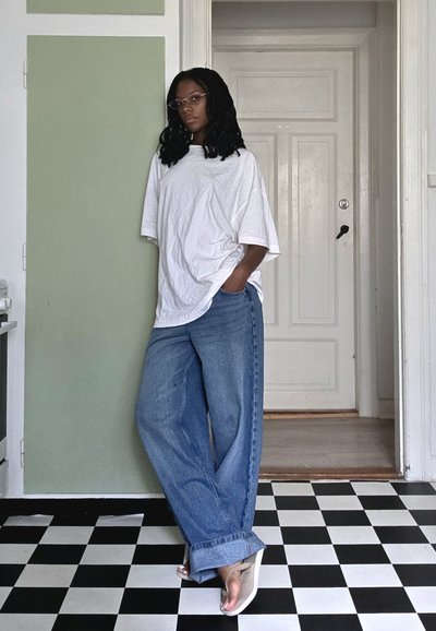 Camiseta blanca de corte holgado, jeans de mezclilla azul claro de corte amplio y sandalias beige. Suelo de cuadros en blanco y negro y pared verde de fondo.