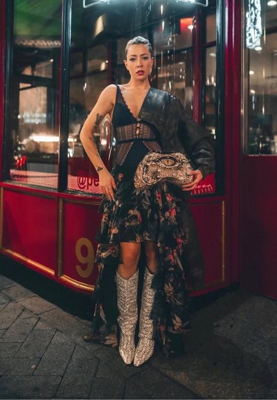 Joven con vestido negro de encaje y falda floral sostiene un bolso de mano con estampado de reptil, lleva botas de piel de serpiente y una chaqueta negra sobre un hombro.
