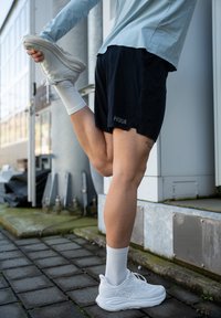 Melnas sporta šorti, gaismzilas krāsas krekls ar garām piedurknēm, baltas sporta apavi ar teksturētu zoli un baltas potīšu zeķes. Izstiepj vienu kāju atpakaļ.