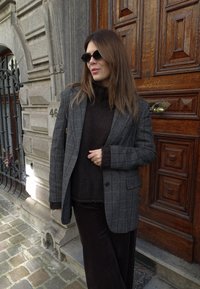 Grå rutete blazer over en brun strikkegenser og mørkebrune bukser. Runde svarte solbriller. Tre dør med dekorative detaljer i bakgrunnen.