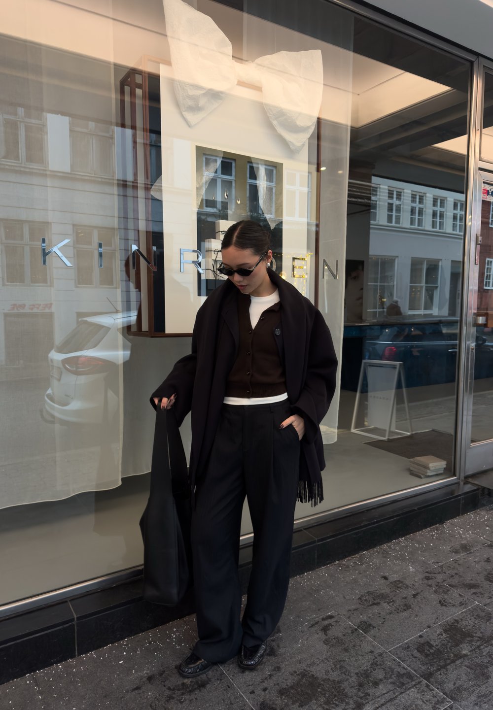 Un vêtement extérieur noir tombe sur un cardigan marron, associé à un pantalon noir plissé à taille haute et un sac cabas noir. Des lunettes de soleil complètent le look.