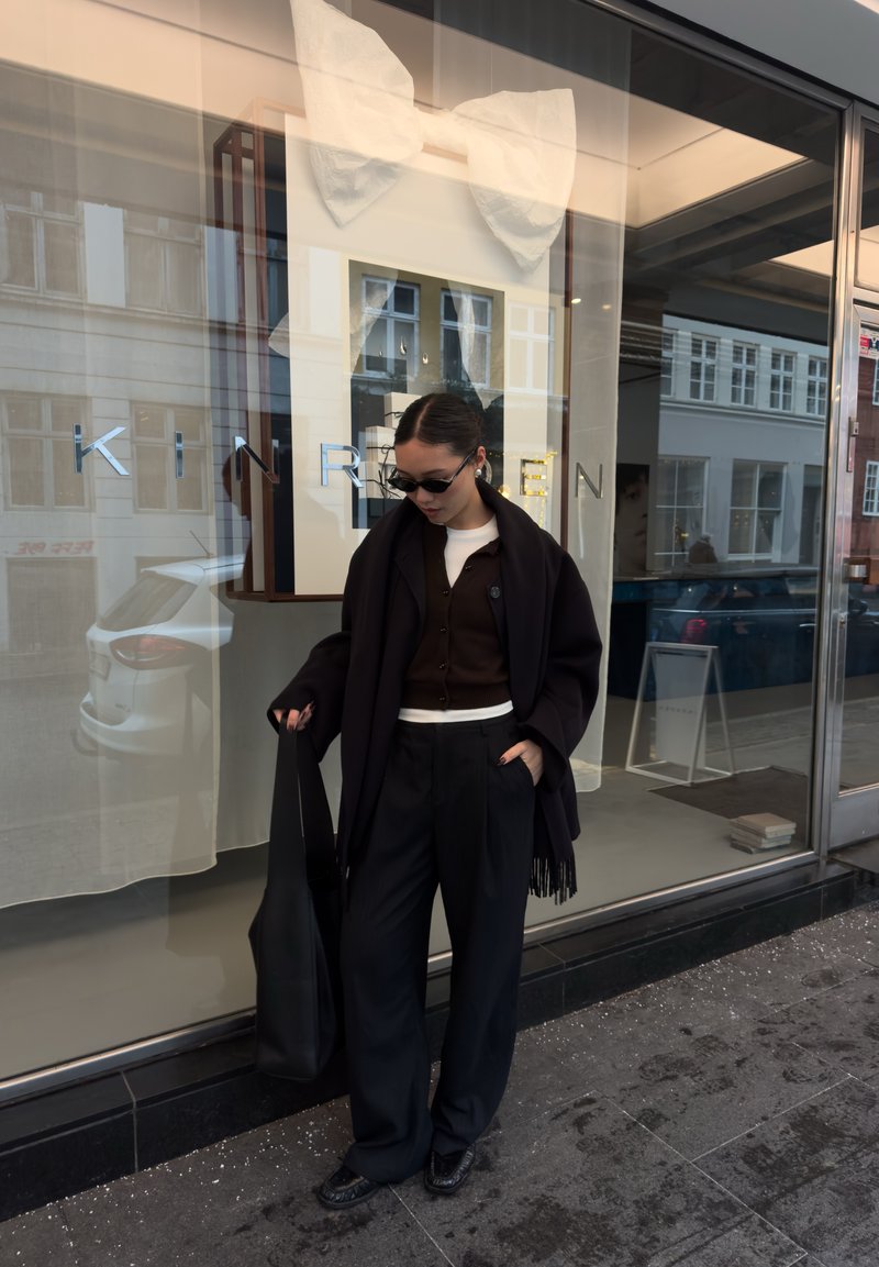 Un vêtement extérieur noir tombe sur un cardigan marron, associé à un pantalon noir plissé à taille haute et un sac cabas noir. Des lunettes de soleil complètent le look.