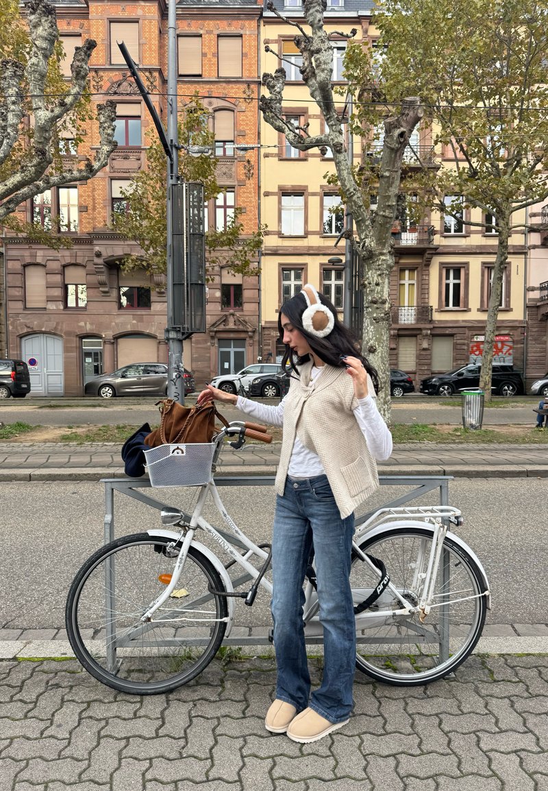 Bicicleta branca com um cesto de arame, jeans azuis desbotados, colete de malha creme e chinelos bege. Protetores auriculares macios apresentam detalhes em pele castanha.