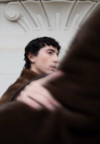 Person med kort krøllet sort hår iført en brun pelsjakke, der holder et stykke stof tæt, med en dekorativ hvid arkitektonisk baggrund.