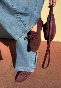 Chaussures en daim bordeaux avec lacets et œillets argentés, associées à un jean bleu clair à jambes larges et un sac bordeaux assorti.