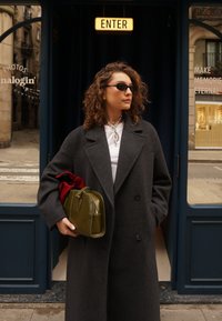 Manteau en laine gris, chemise blanche, sac en cuir vert avec une fermeture éclair, écharpe rouge et lunettes de soleil. En arrière-plan, l'entrée d'un magasin avec des moulures bleues.
