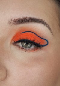 Vivace ombretto arancione con contorno blu, lunghe ciglia nere e un tono di pelle naturale. I dettagli includono sopracciglia ben curate e pelle liscia.