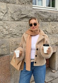 Veste en fausse fourrure beige avec des boutons noirs, pull rose clair, écharpe tricotée beige, gants beiges et jean bleu, tenant des tasses de café et un sac à main.