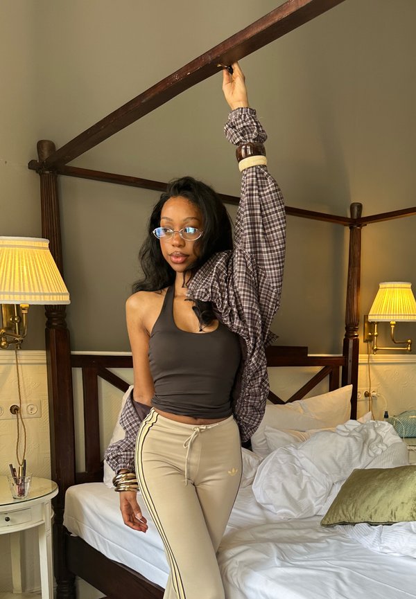 Ung kvinde med briller og ternet skjorte over den ene arm står ved siden af en uopredt seng og holder i en træbeklædt baldakin med den ene hånd.