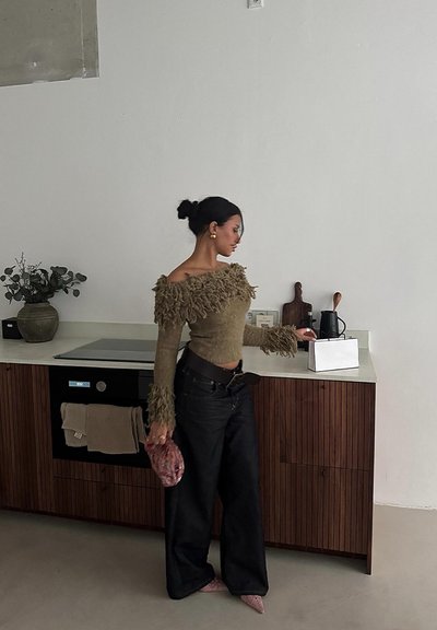 Suéter verde oliva sin mangas con acentos de flecos, jeans negros de pierna ancha, tacones rosas y un bolso estampado en un ambiente de cocina moderna.