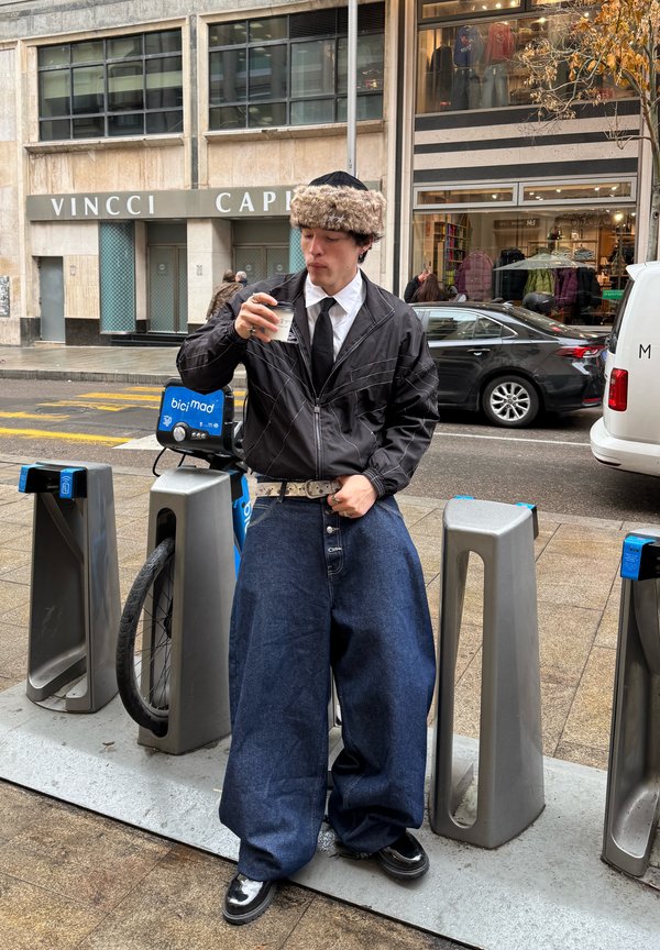 Mann iført vide mørke jeans, svart jakke, pelslue og slips, står ved en sykkeldelingsstasjon og drikker kaffe på en bygate.