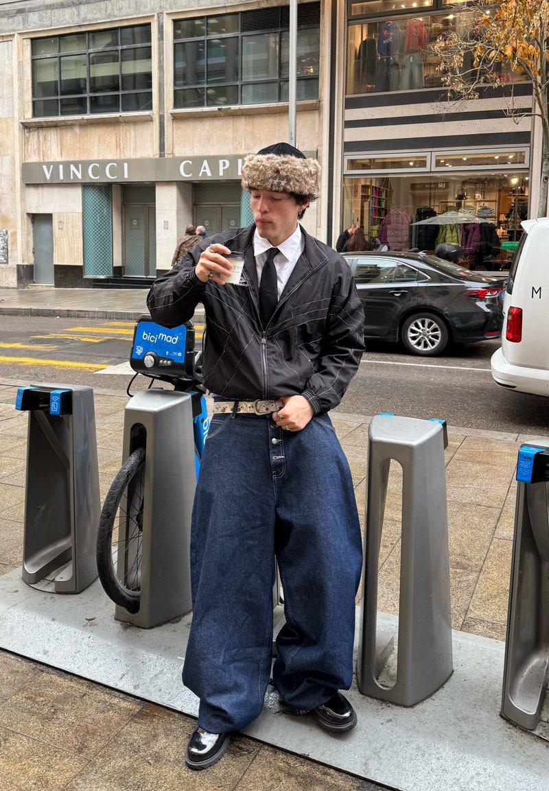 Muškarac u širokim tamnim trapericama, crnoj jakni, krznenoj kapi i kravati stoji pored stanice za dijeljenje bicikala, pijući kavu na ulici u gradu.
