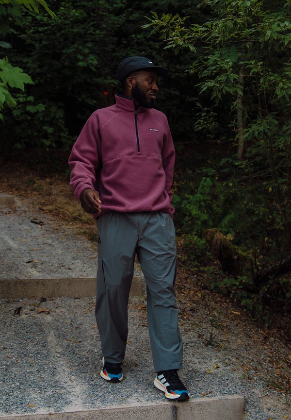 Man draagt een paarse fleece, grijze broek en sneakers, loopt een stenen trap af omgeven door dichte, groene bomen en begroeiing.