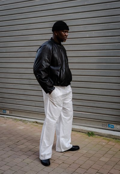 Chaqueta bomber de cuero negra, pantalones de pierna ancha blancos y zapatos negros. De pie contra un fondo gris, con las manos en los bolsillos y expresión neutral.