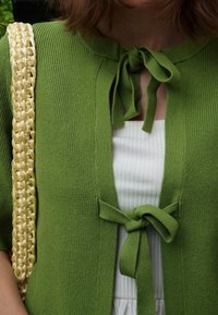 Vihreä kardigan, jossa on ribbikuvio, etuosassa solmimisella kiinnitys. Päällä valkoinen ribbikuvioinen paita, jota täydentää keltainen punottu olkalaukku.