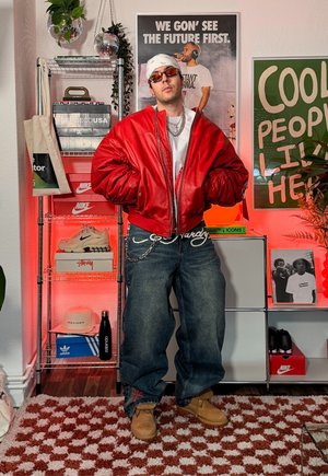 Hombre con chaqueta roja, bandana y gafas de sol de pie en una habitación con estanterías de zapatillas y pósters que dicen "Primero veremos el futuro" y "Aquí viven personas geniales."
