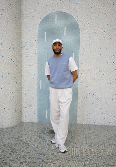 Sudadera sin mangas azul claro sobre una camiseta blanca, combinada con pantalones blancos y zapatillas, de pie contra un fondo azul de azulejos.