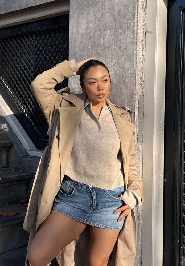 Beige trench coat over a grey sweater and denim mini skirt. Textured fabric, relaxed fit, short sleeves, and a neutral color palette.