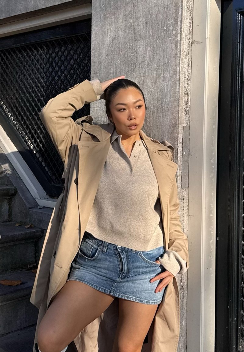 Trench beige sopra un maglione grigio e una minigonna di jeans. Tessuto strutturato, vestibilità comoda, maniche corte e una palette di colori neutri.