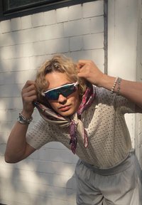 Kék és lila lencséjű napszemüveg, mintás selyemsál a nyak körül, világos kötött pulóver, szürke sort és ezüst karkötő.