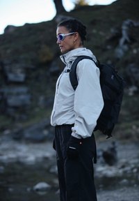 Vrouw met zonnebril, lichte jas, zwarte handschoenen en rugzak, staand buiten op rotsachtig terrein, in profiel naar links kijkend.