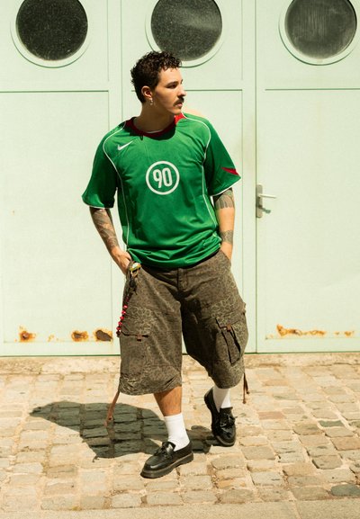 Jersey verde de Nike con detalles rojos y logo circular "90", emparejado con pantalones cortos de carga khaki texturizados y zapatos negros, de pie sobre adoquines.