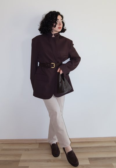 Mujer con cabello rizado, lleva un blazer oscuro de gran tamaño ceñido a la cintura, pantalones beige, mocasines oscuros, sostiene un bolso negro, de pie sobre suelo de madera.