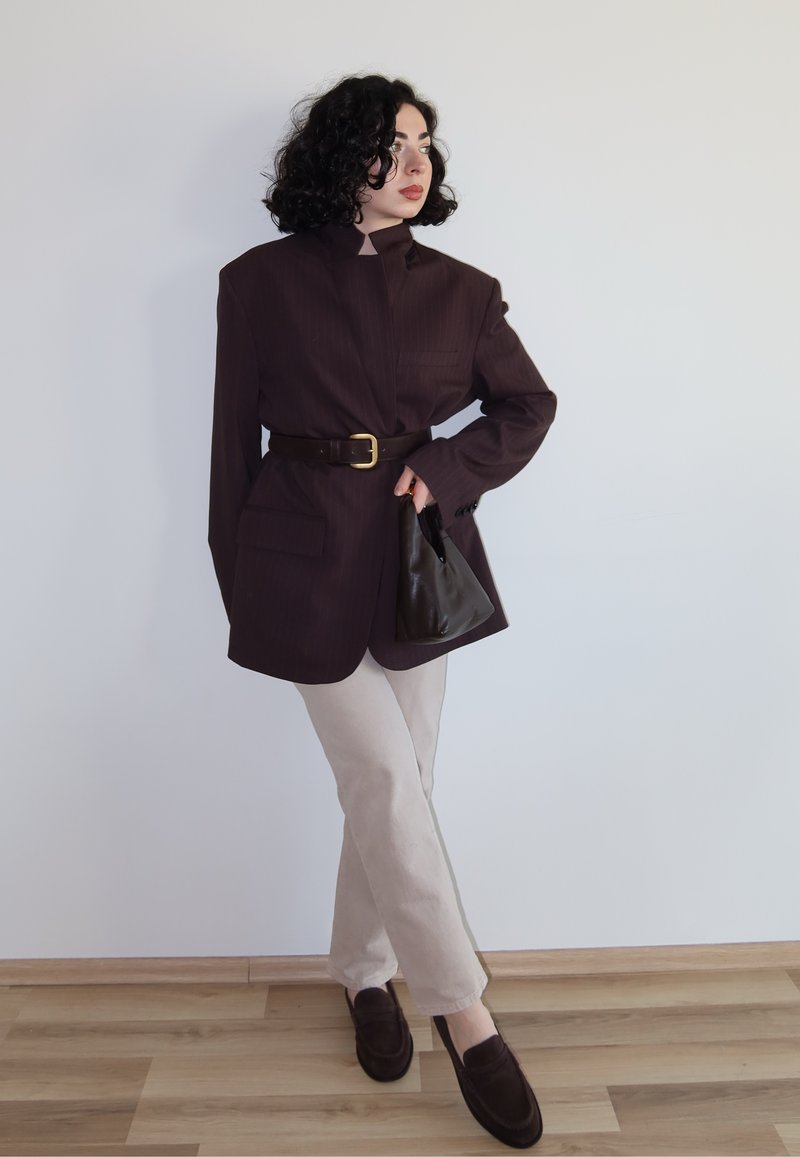 Žena kovrčave kose koja nosi preveliki tamni blejzer s remenom u struku, bež hlače, tamne loaferice, drži crnu torbu, stoji na drvenom podu.