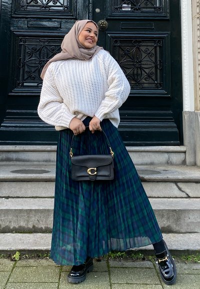 Suéter de punto crema, falda plisada de cuadros verde oscuro, bolso negro con herrajes dorados y mocasines negros brillantes. De pie en escalones de piedra.