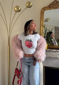 White cropped t-shirt featuring a large strawberry graphic, paired with a light pink fluffy jacket. Light blue wide-leg jeans and a red handbag.