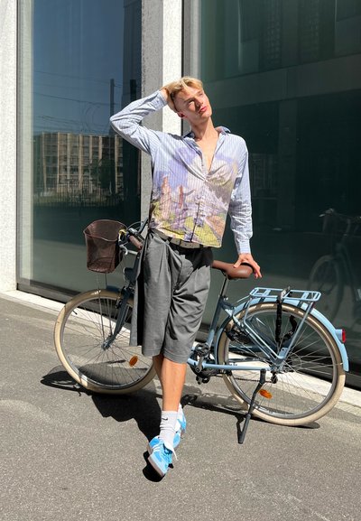 Hombre vistiendo una camisa a rayas con un estampado de paisaje, pantalones cortos grises y zapatillas azules, de pie al lado de una bicicleta azul clara con una cesta marrón.