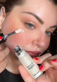 Luminous Silk -korostusvoide lasipullossa, jossa hopeanvärinen korkki; kermaisen valkoinen tuote levitettynä iholle siveltimellä. Kirkkaat kynnet näkyvissä.