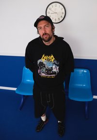 Homem com barba e boné, usando um hoodie gráfico preto, está entre cadeiras azuis de sala de espera, sob um relógio de parede que marca 9:42.