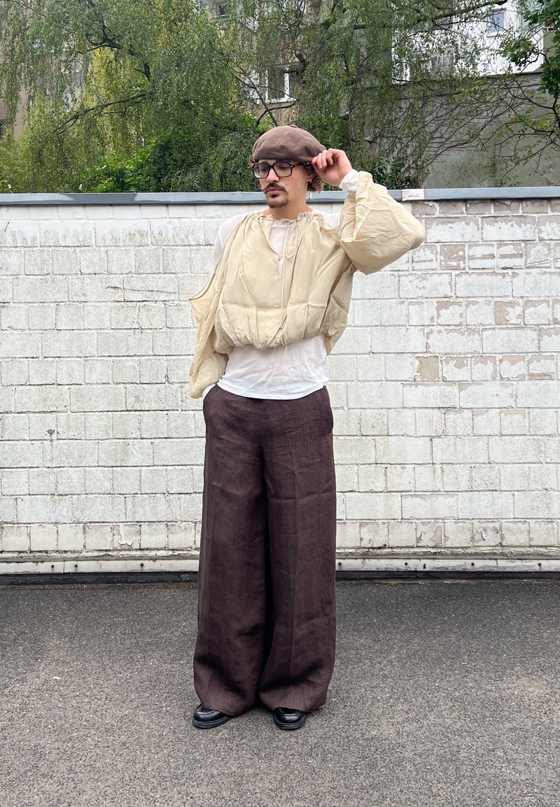 Personne portant une veste beige matelassée sur une chemise blanche, un pantalon large marron, des chaussures noires et un béret marron, debout sur l'asphalte près d'un mur en briques blanches.