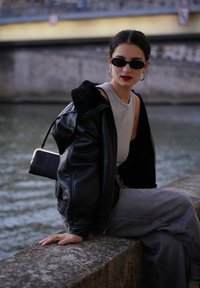 Schwarze Lederjacke mit einem flauschigen Kragen, graues ärmelloses Oberteil und graue weit geschnittene Hose; schwarze rechteckige Handtasche; minimalistische schwarze Sonnenbrille.