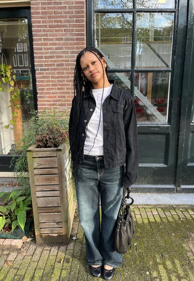 Joven con trenzas largas se apoya ligeramente, lleva una chaqueta negra, una camisa blanca, vaqueros azules y sostiene un bolso negro junto a una pared de ladrillos y una puerta de cristal.