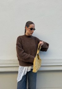 Moteris su nupinta šukuosena, dėvinti tamsius akinius nuo saulės, rudą megztinį, baltą nėriniuotą viršūnę ir džinsus, laiko geltoną studuotą rankinę šalia šviesios sienos.