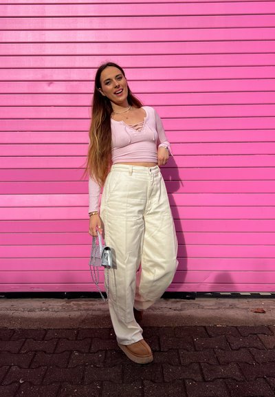 Top de manga larga rosa con escote de encaje combinado con pantalones anchos blancos. Sosteniendo un pequeño bolso de mano plateado con cadena. Fondo rosa.