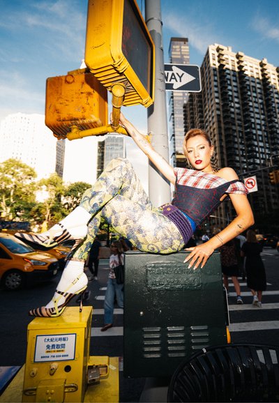 Modelo vestida con una blusa de cuadro sin mangas, pantalones con estampado y sandalias de tacón alto con tiras, sentada sobre una caja de control de tráfico verde.