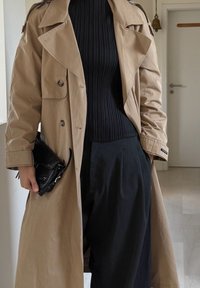 Trench-coat beige avec une ceinture à la taille, des boutons croisés et des épaulettes, associé à un haut noir côtelé et un pantalon noir ample.