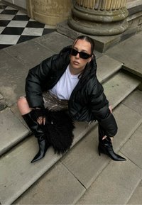 Musta kiiltävä toppatakki, valkoinen T-paita, kuviolliset shortsit, polven korkeuteen ulottuvat kärjelliset mustat saappaat ja pörröinen musta käsilaukku.