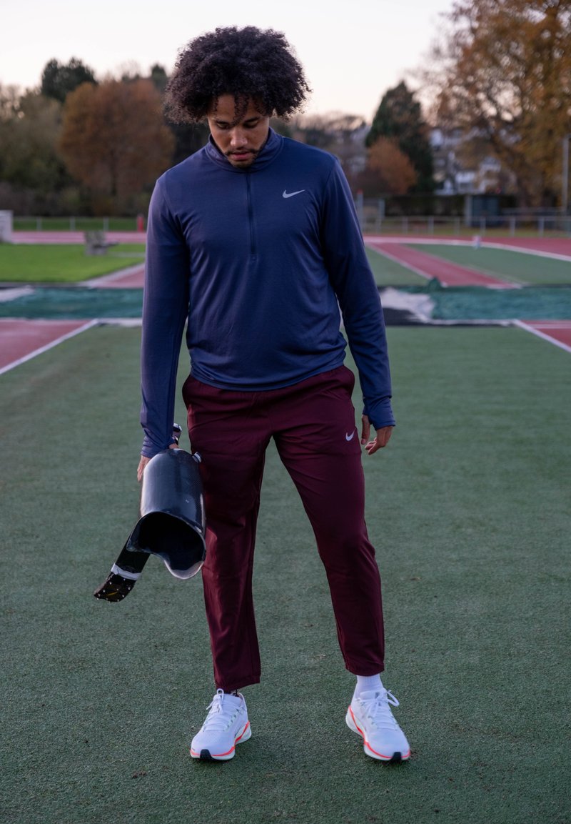 Homem em roupa desportiva segurando uma perna protética, em pé num campo desportivo ao ar livre durante o outono.
