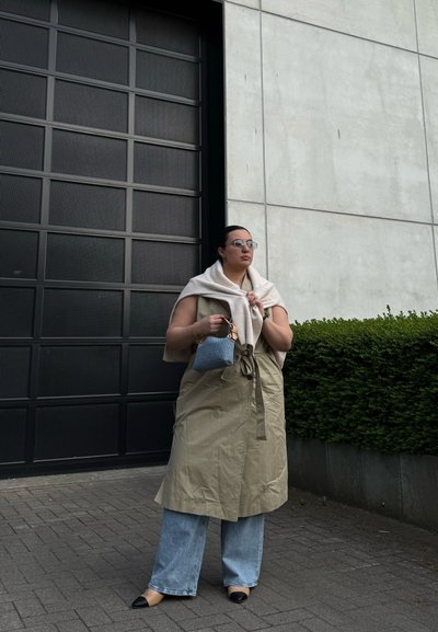 Gabardina beige con cinturón, abrigo claro, bolso azul, jeans cortos claros y zapatos negro y marrón. Puerta de garaje gris en el fondo.