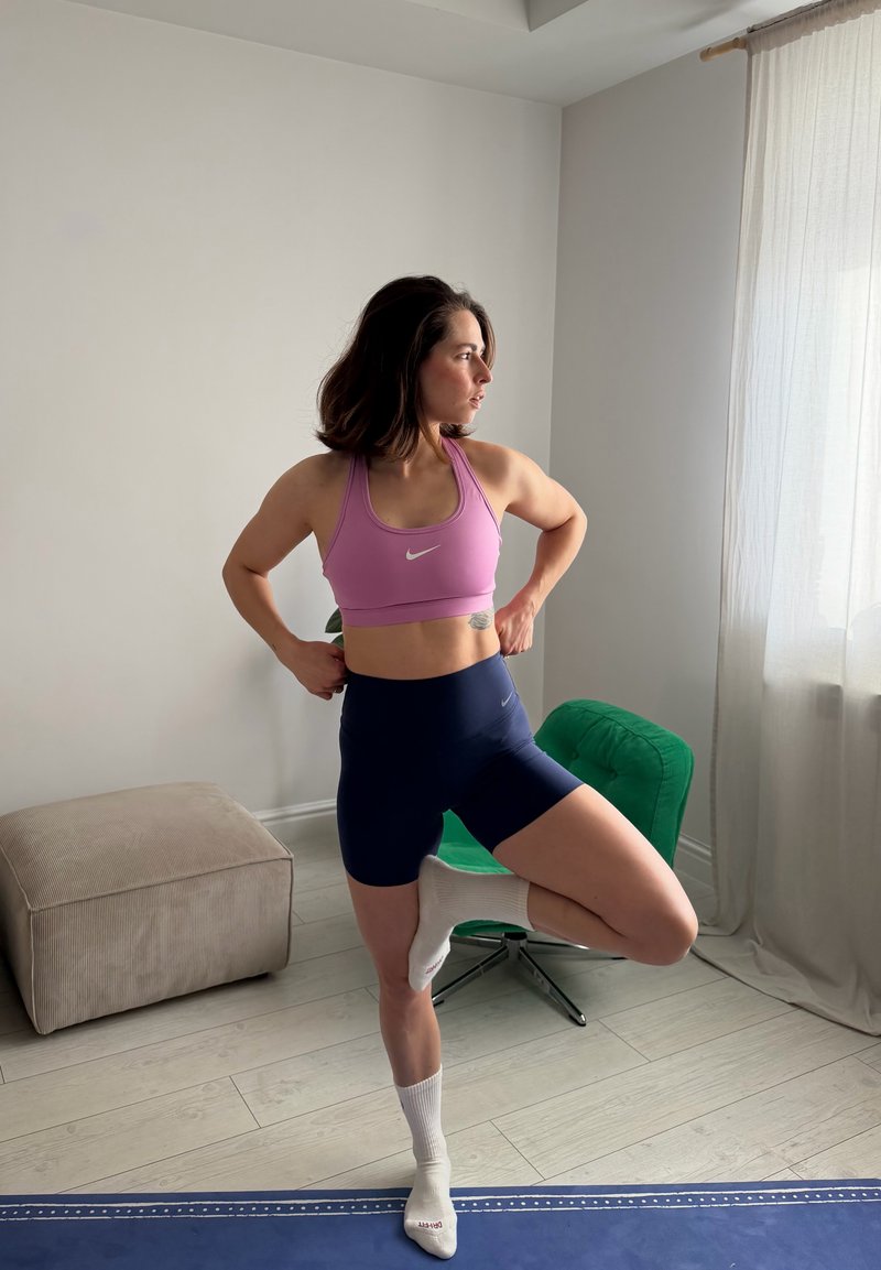 Kvinna i rosa sport-bh och marinblå shorts balanserar på ett ben på en blå yogamatta i ett ljust rum med beige och gröna möbler.
