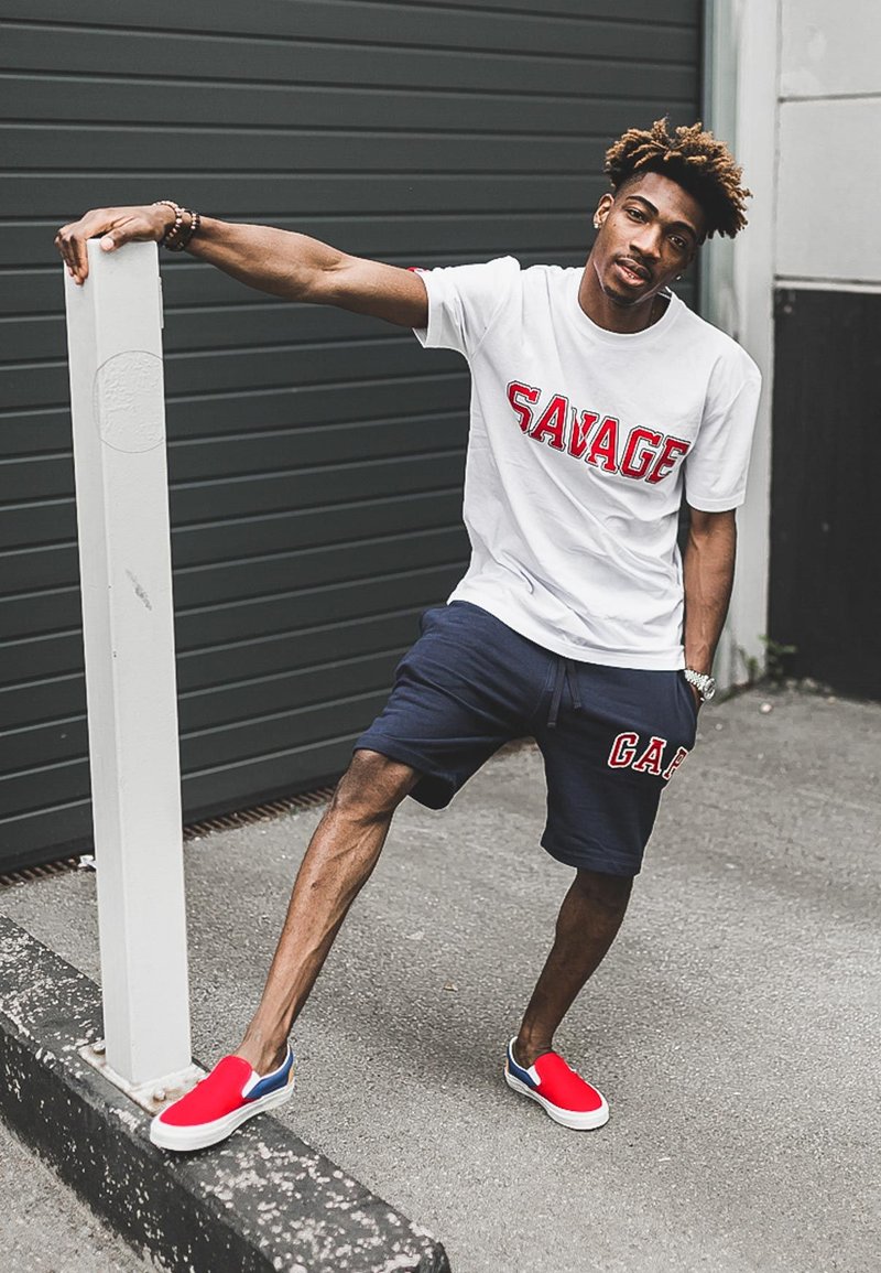 White t-shirt featuring "SAVAGE" in red letters, navy shorts with "GAP" branding, red slip-on shoes, posed against a grey background.