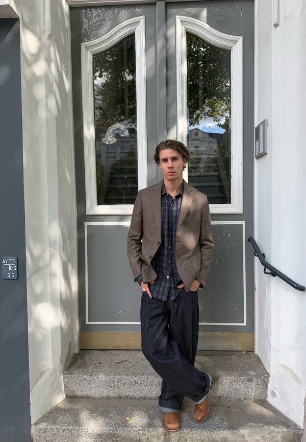 Young man in casual blazer and plaid shirt leaning against gray door with glass panels on stone steps outdoors.