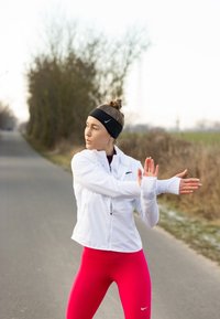 Veste de sport blanche à fermeture éclair devant, avec des accents noirs, portée avec des leggings roses ajustés. Un bandeau noir complète le look de sport.