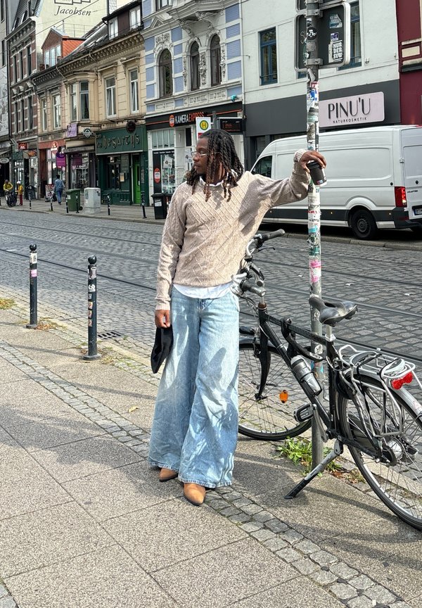 Maglione a maglia color beige chiaro con motivo a rombi, abbinato a jeans in denim blu sbiadito a gamba larga. Scarpe marroni e un accessorio nero in mano.
