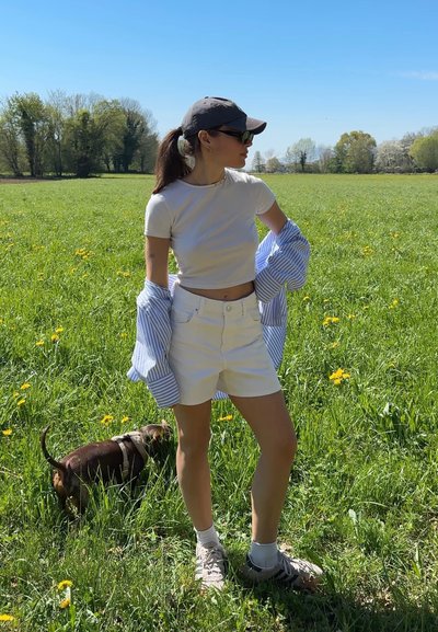 Mujer con pantalones cortos blancos y top corto de pie en un campo de hierba con flores amarillas, lleva gorra y gafas de sol, con un pequeño perro marrón cerca.